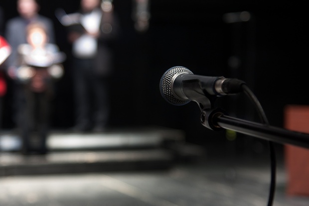 En mikrofon i fokus på en scen med suddiga personer i bakgrunden, troligen en kör eller grupp som förbereder sig.