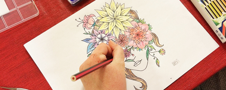 En person färglägger en teckning av ett ansikte med blommor i håret vid ett bord med färgpennor och pärlor.