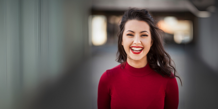 Leende kvinna med långt mörkt hår och röd tröja står utomhus med suddig bakgrund.