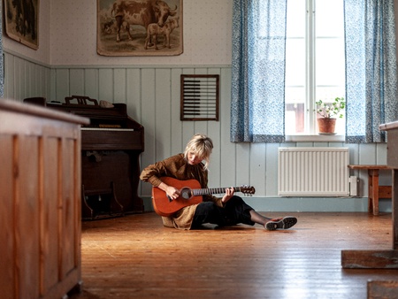 Person sitter på ett trägolv i ett klassrum och spelar akustisk gitarr, med ett piano i bakgrunden.