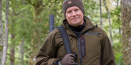 Man i brun jacka och mössa står i skog med gevär på axeln och ler mot kameran.