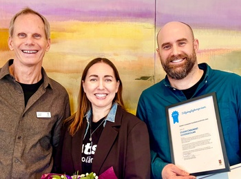 Tre personer står framför en färgglad bakgrund, håller blommor, diplom och en stor prischeck.