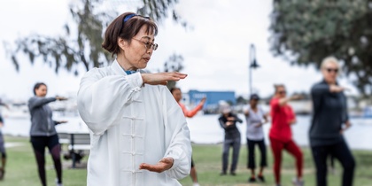 En grupp människor utövar tai chi utomhus i en park, med en kvinna i vit dräkt i förgrunden.