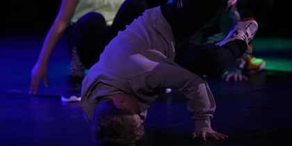 En person utför ett breakdance-move på golvet med stöd av ena handen, andra personer i bakgrunden.