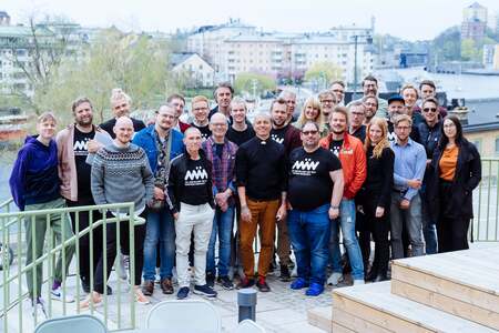 En grupp människor står tillsammans utomhus på en terrass med stad och vatten i bakgrunden.