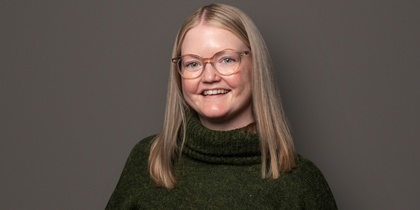 Person med långt blont hår, grön stickad tröja och svarta byxor står mot en grå bakgrund.