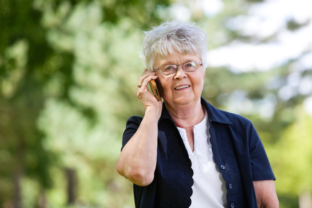 Äldre kvinna med kort grått hår pratar i mobiltelefon utomhus i en grön park.