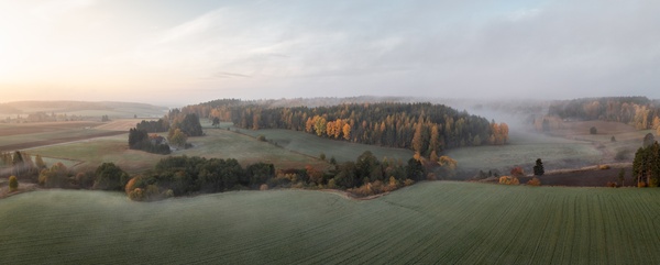 Panoramabild över ett dimmigt landskap med åkrar, skog och morgonljus.