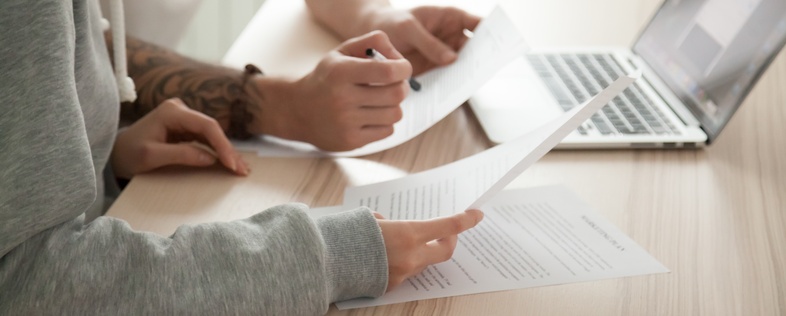 Två personer sitter vid ett bord och granskar dokument framför en bärbar dator.