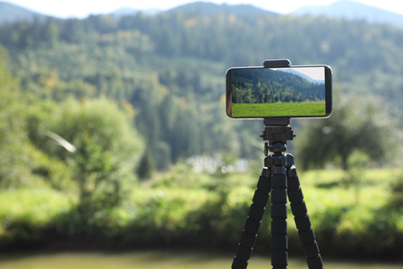 En smartphone på stativ fotograferar ett grönt landskap med skog och berg i bakgrunden.
