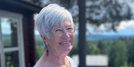 Lena Söderlund står utomhus i solsken, bär vit klänning och röda örhängen, ler mot kameran.