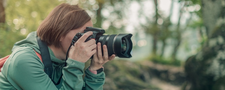 Person med kamera fotograferar i en skogsmiljö, klädd i grön jacka och ryggsäck.