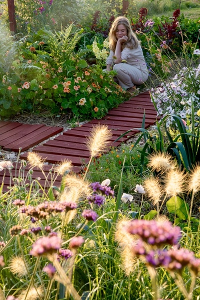 En kvinna sitter på huk vid en blomrabatt längs en röd trästig i en solig trädgård.