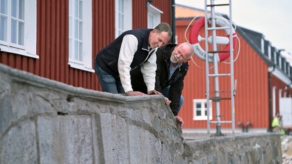 Två män lutar sig över en stenmur och skrattar utanför en röd träbyggnad.