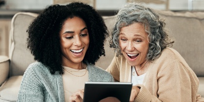 Två kvinnor sitter på golvet framför en soffa och tittar glatt på en surfplatta tillsammans.