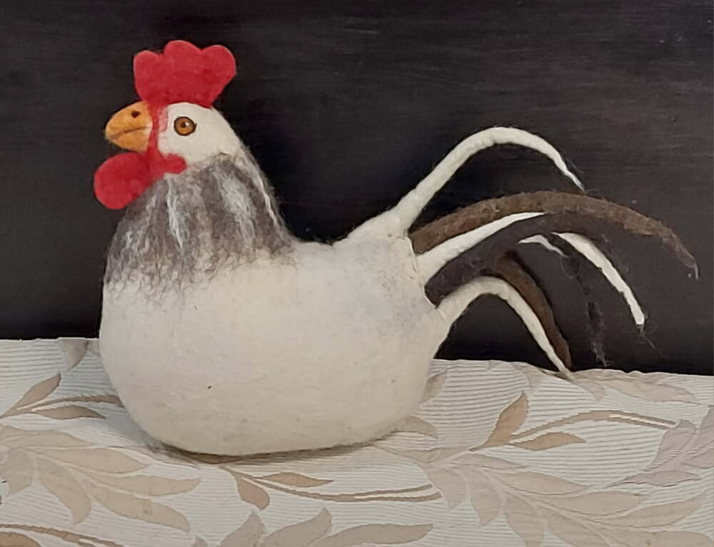 En filtad tupp i vitt, rött och brunt står på ett mönstrat tyg framför en mörk bakgrund.