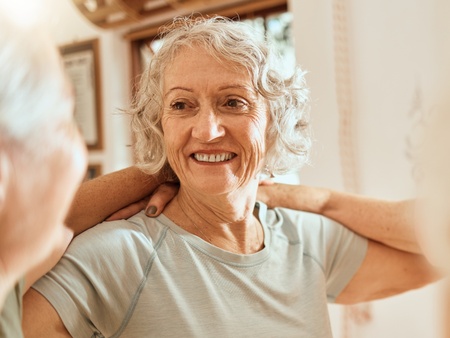 Äldre kvinna med grått hår ler och gör stretchövningar inomhus.