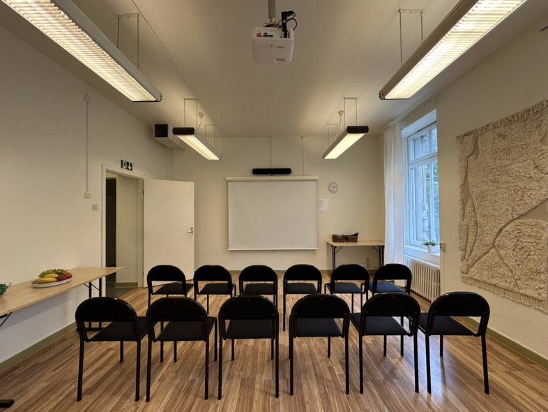 Ett tomt klassrum med stolar i rader, projektorduk, projektor i taket och stora fönster.