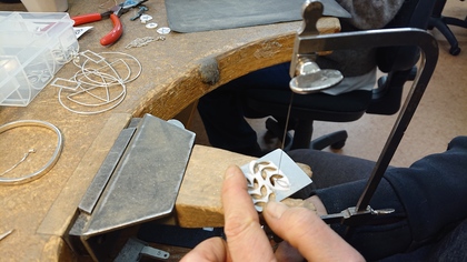 En person sågar i en metallplatta vid ett arbetsbord, omgiven av smyckesverktyg och material.