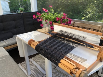 En vävstol med svart tyg står på ett bord i ett ljust uterum med blommor och sittplatser.