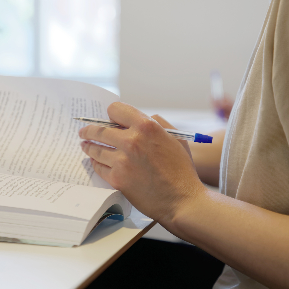 En person sitter vid ett bord och läser en bok, håller en penna i handen.