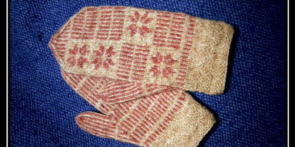 Ett par stickade vantar med röd och beige mönster ligger på ett blått tygunderlag.