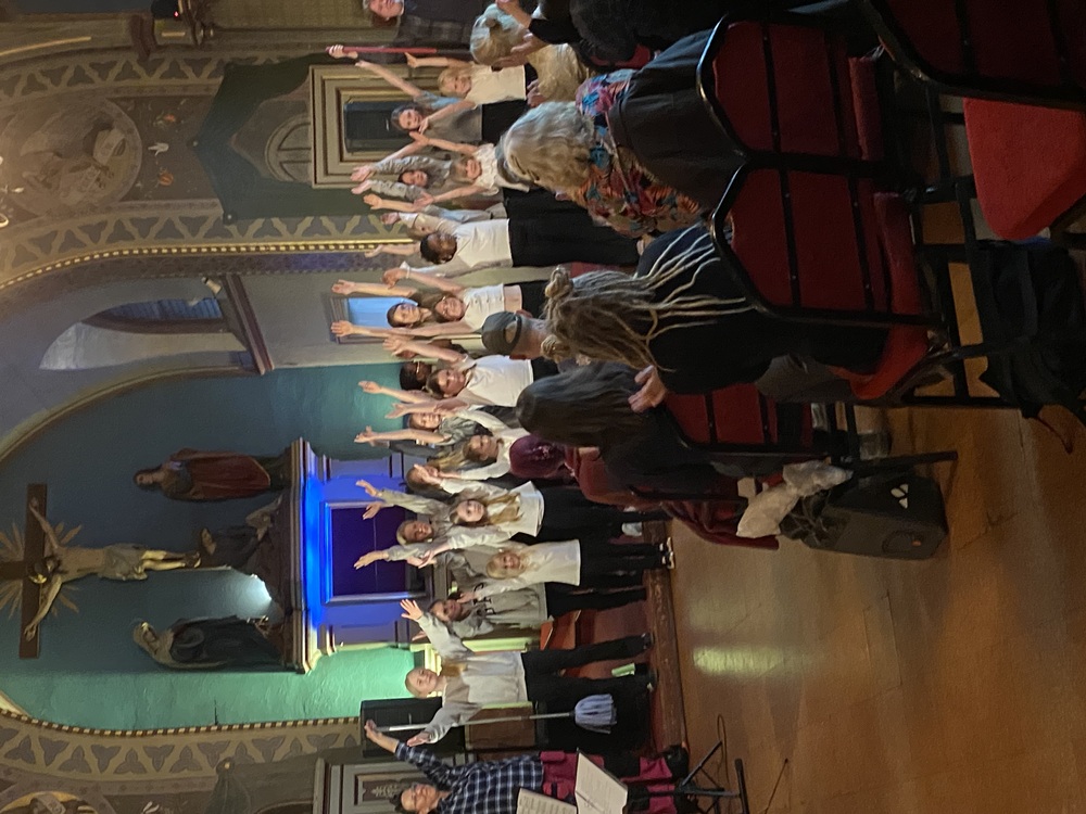 En barnkör står på scen i en kyrka med händerna i luften inför en sittande publik.