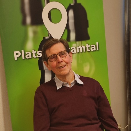 Man med glasögon sitter vid ett bord framför en grön skylt med texten 'Plats för samtal'.