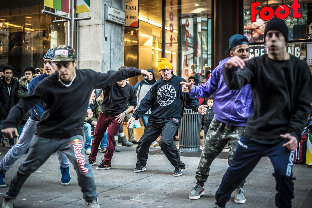 En grupp unga män dansar hiphop på en stadsgata, omgivna av åskådare och butiker.