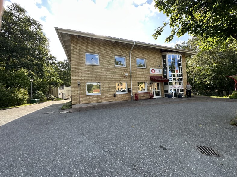 Gul tegelbyggnad med glasentré, omgiven av träd och asfalt, en person står utanför.