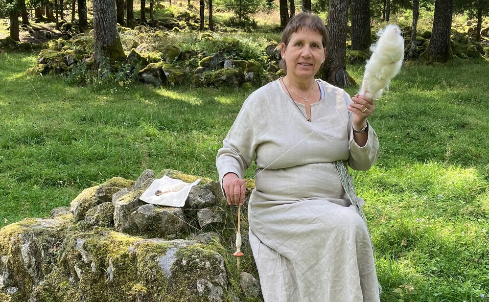 Kvinna i ljus klänning sitter på en mossig sten i skog och spinner ull med slända.