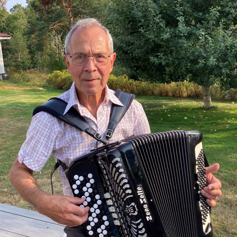 Äldre man spelar dragspel utomhus på en altan med träd och gräs i bakgrunden.