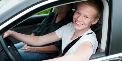 Ung man ler och sitter bakom ratten i en bil, med en vuxen passagerare bredvid sig.