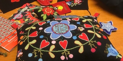 Två personer arbetar med färgglad broderi på svart tyg vid ett bord, med kuddar och tygbitar.