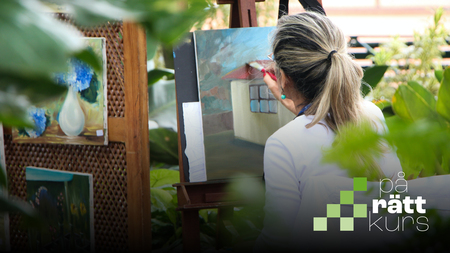 Kvinna målar en tavla utomhus omgiven av växter och andra konstverk.