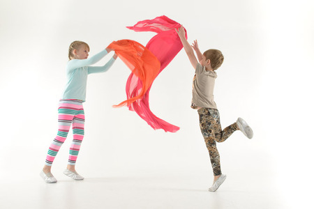 Två barn leker med färgglada tygsjalar i ett ljust rum. Ett barn hoppar medan det andra håller i sjalen.