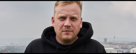 En man med blont hår och svart hoodie står utomhus med en stad i bakgrunden under mulen himmel.