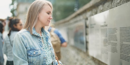 Kvinna i jeansjacka läser informationstavla utomhus vid en stenmur.