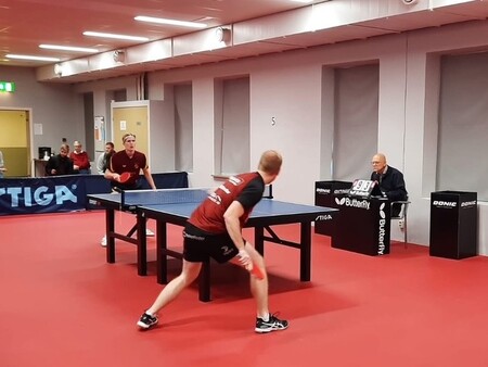 Två män spelar bordtennis i en inomhushall medan en domare räknar poäng vid sidan av bordet.