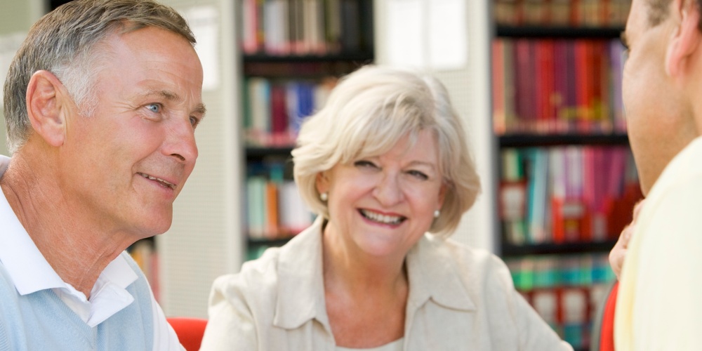 Tre äldre vuxna sitter vid ett bord i ett bibliotek och samtalar.