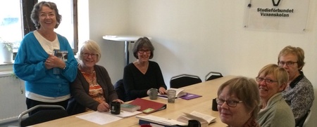 Sju äldre kvinnor sitter runt ett bord i ett mötesrum med papper, böcker och kaffekoppar.