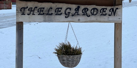 Träskylt med texten 'THELEGÅRDEN' under ett snötäckt tegeltak, med en hängande korg och snö på marken.