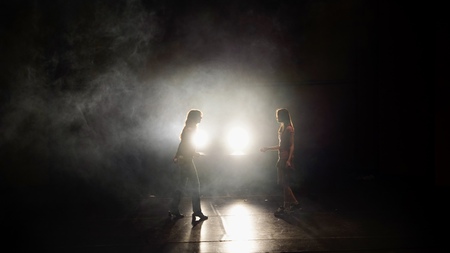 Två personer står mittemot varandra på en mörk scen med starkt motljus och rök.