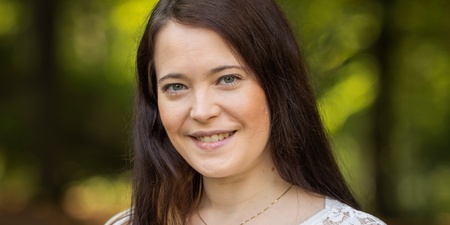 Kvinna med långt brunt hår och vit spetsblus står utomhus och ler mot kameran, grönskande bakgrund.