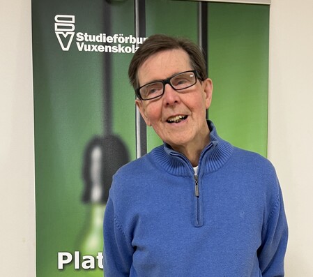 Person i blå tröja står framför en grön rollup med texten 'Studieförbundet Vuxenskolan'.