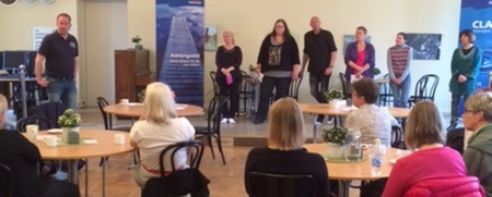 Ett antal personer sitter vid runda bord i en ljus sal, medan några står framför och verkar presentera något.