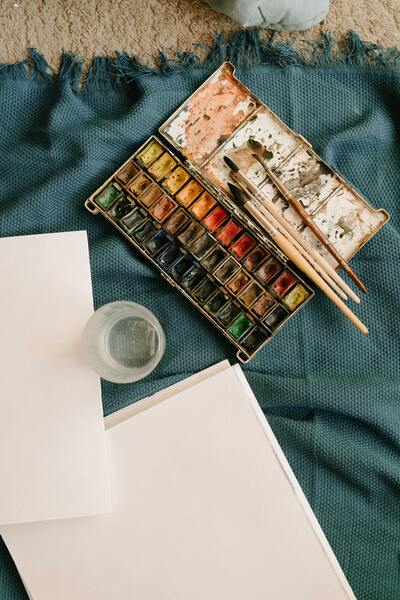 Akvarellfärger, penslar, papper och ett glas vatten ligger på en blå filt, redo för målning.