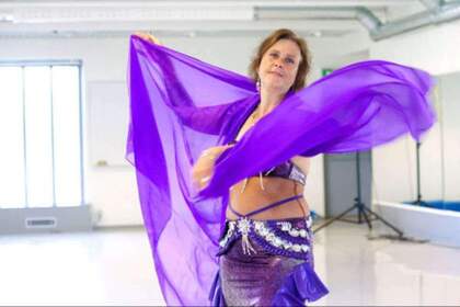 Kvinna dansar med lila slöja i ett ljust rum, klädd i glittrande magdansdräkt.