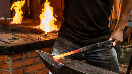 En person smider ett glödande järn på ett städ med eld i bakgrunden.