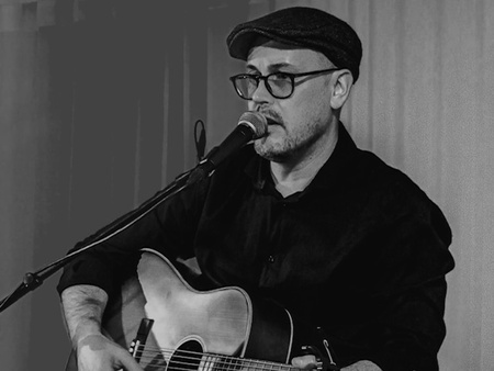 En man med glasögon och basker som sitter på en stol och spelar gitarr samt sjunger i en mikrofon. Svartvit bild.